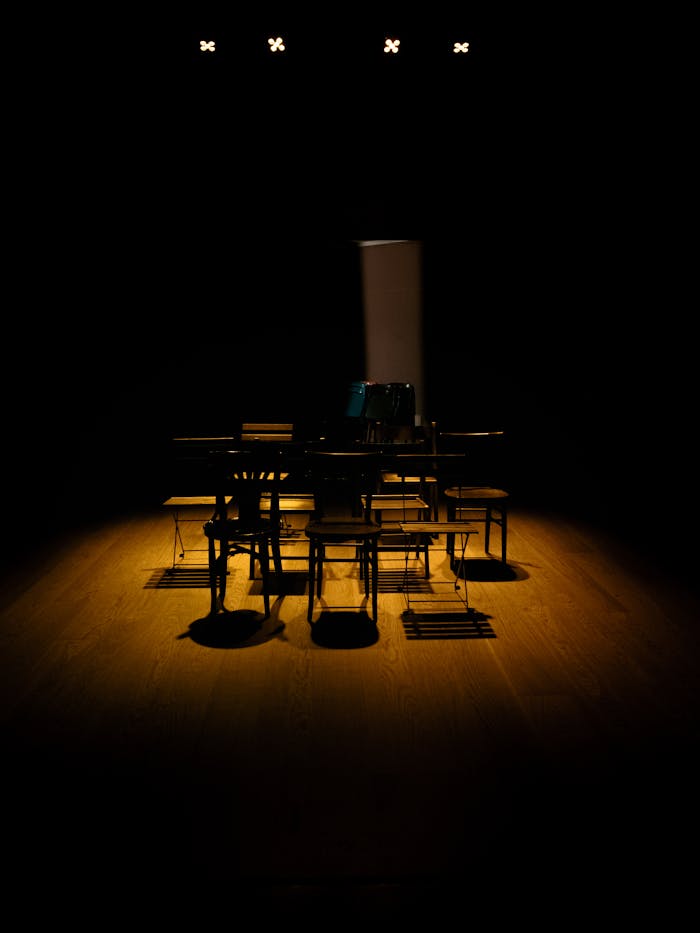 Dark theater stage with chairs and a single light focus, creating dramatic shadows.
