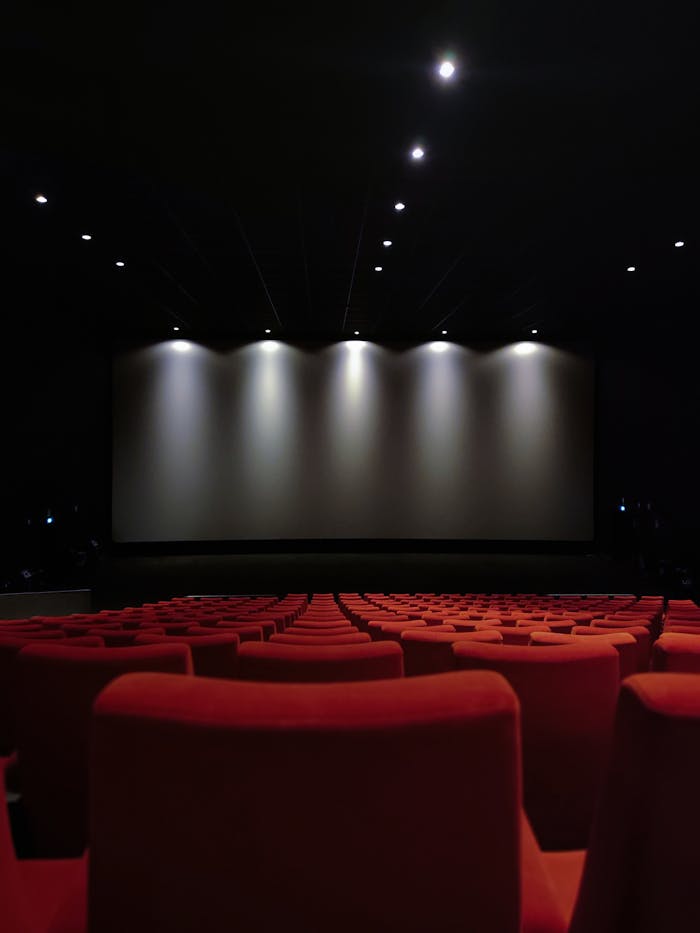 View of an empty cinema with rows of red seats and a large screen illuminated by lights.